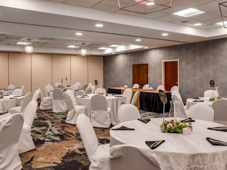 The image shows a banquet hall with round tables covered in white cloths, black napkins, floral centerpieces, and chairs, set up for an event.