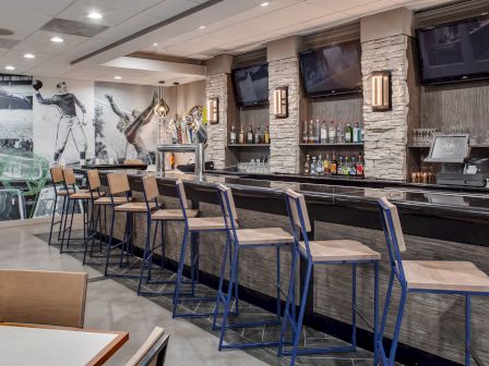 This image shows a modern bar with high stools, a row of liquor bottles, TV screens, and sports-themed wall art. The setting is sleek and inviting.