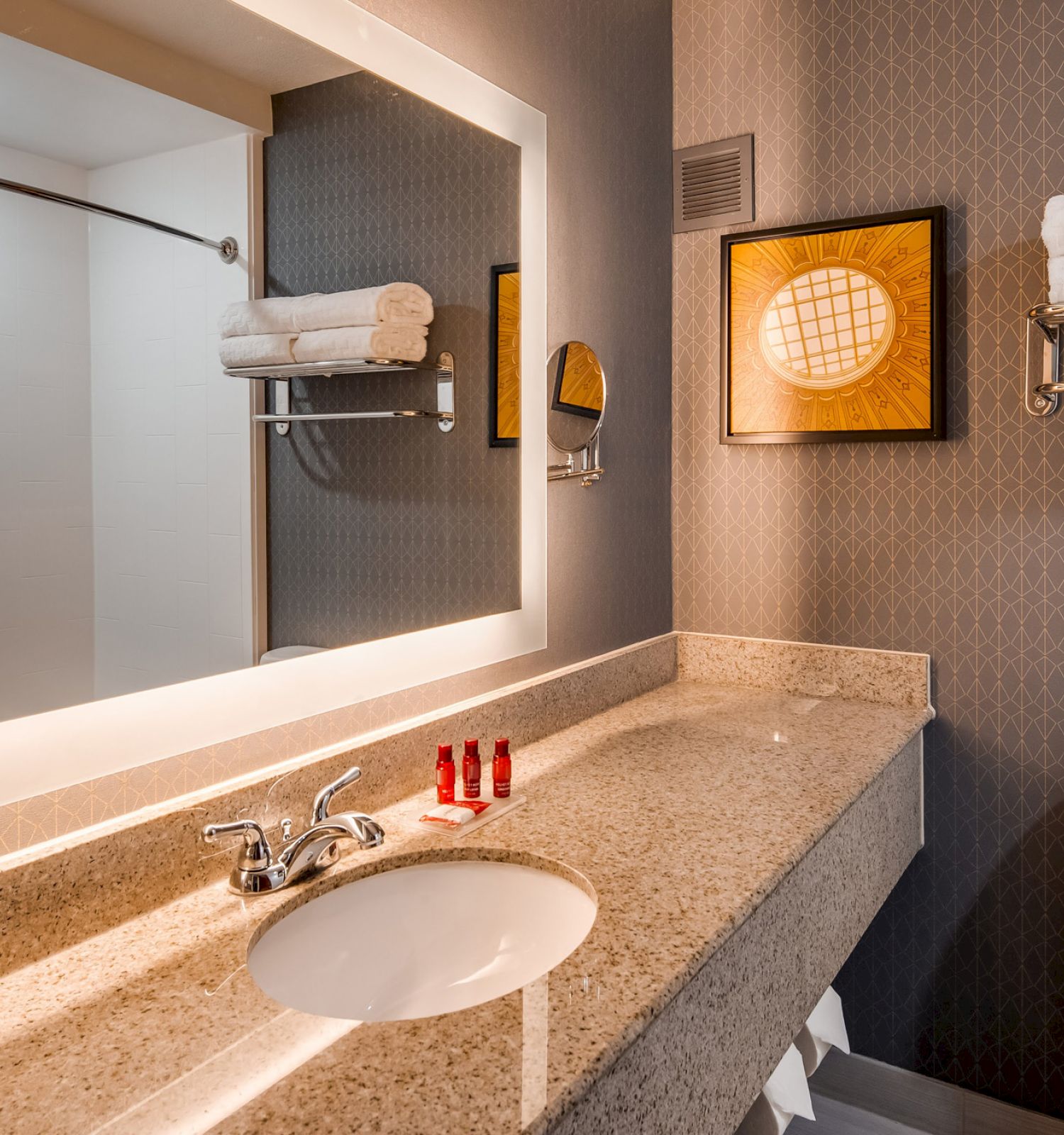 A modern bathroom featuring a large illuminated mirror, sink with red toiletries, toilet, towel rack with folded towels, and framed artwork.