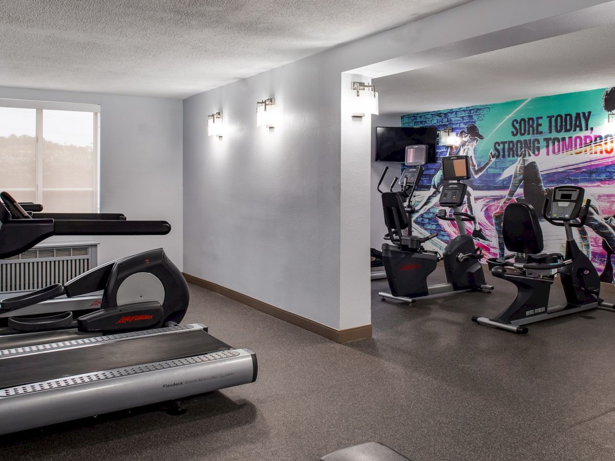 The image shows a gym with treadmills, exercise bikes, and a motivational mural that reads 