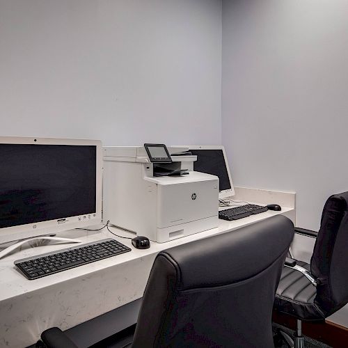 This image shows a small office space with two computer setups, a printer, and two black office chairs against a white wall on a white desk.