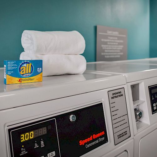 The image shows a laundry room with washing machines and dryers. On top of the machines are a box of detergent and two white towels.