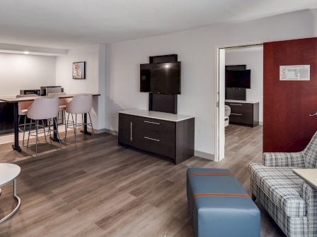 The image depicts a modern hotel room with a kitchenette, dining area, TV, and a seating area. There's a separate room visible through the open door.
