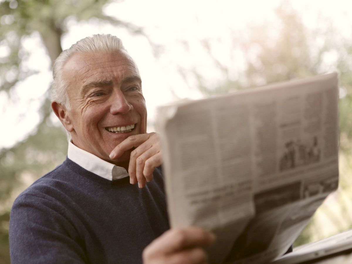 An older man with a bright smile is reading a newspaper outdoors, dressed in a navy sweater and white collared shirt, with trees in the background.
