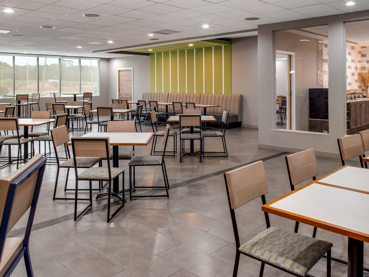 The image features an empty cafeteria with neatly arranged tables and chairs, modern decor, and large windows allowing natural light to enter.