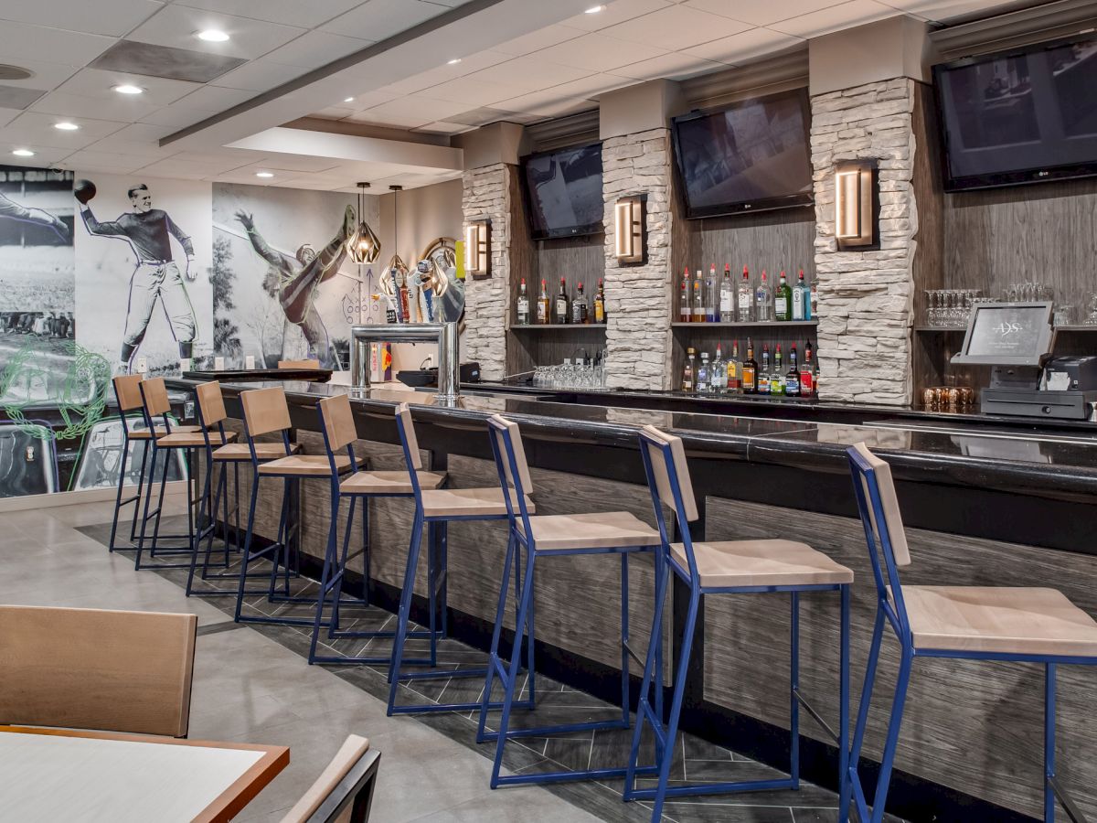 A modern bar with high chairs, stone accents, multiple TVs, a sports mural, and a stocked shelf of spirits, likely in a restaurant or hotel.