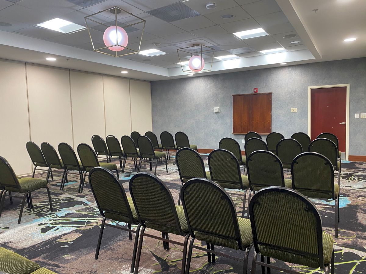 The image shows an empty conference room with rows of green chairs facing a white screen and closed red door, under bright lighting.
