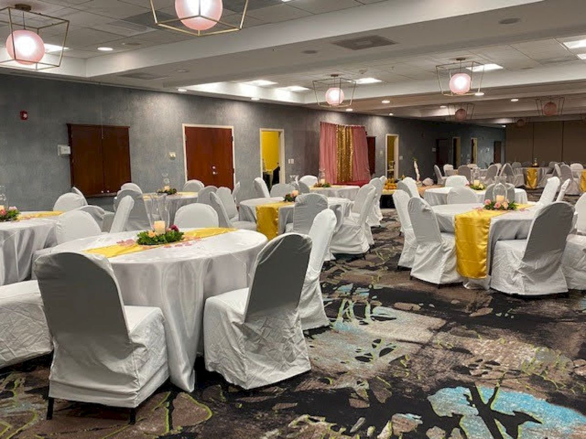 The image shows a decorated banquet hall with round tables covered in white tablecloths and chairs with covers, prepared for an event.