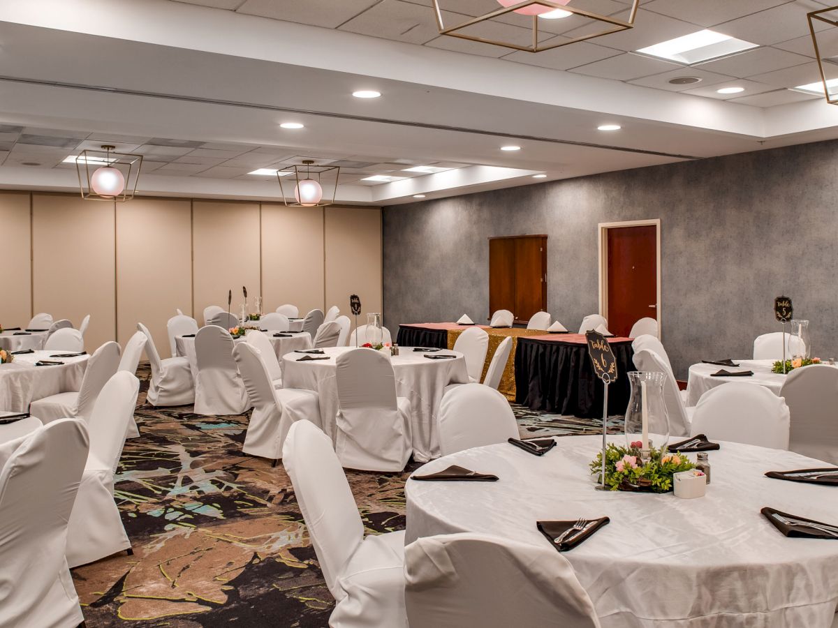 The image shows a banquet hall set up with round tables covered in white tablecloths, each adorned with flower centerpieces and black napkins.