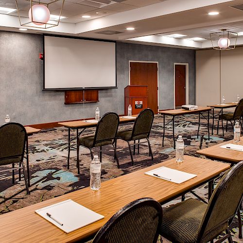 The image shows a conference room setup with tables, chairs, notepads, and water bottles, facing a presentation screen and a podium.