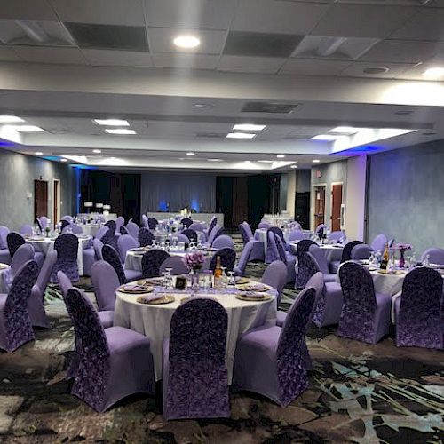 The image shows a banquet hall set up for an event with round tables, purple chair covers, and table settings, ready for guests.