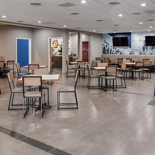 The image shows a modern, empty cafeteria with tables, chairs, a counter area, and televisions on the wall.