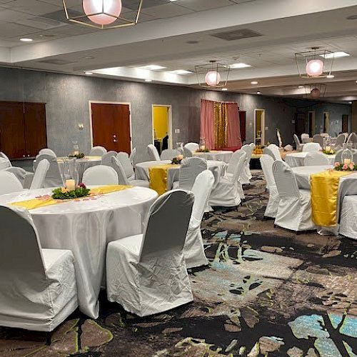 The image shows a spacious event hall with round tables covered in white cloths and chairs draped in white covers, decorated for a function.