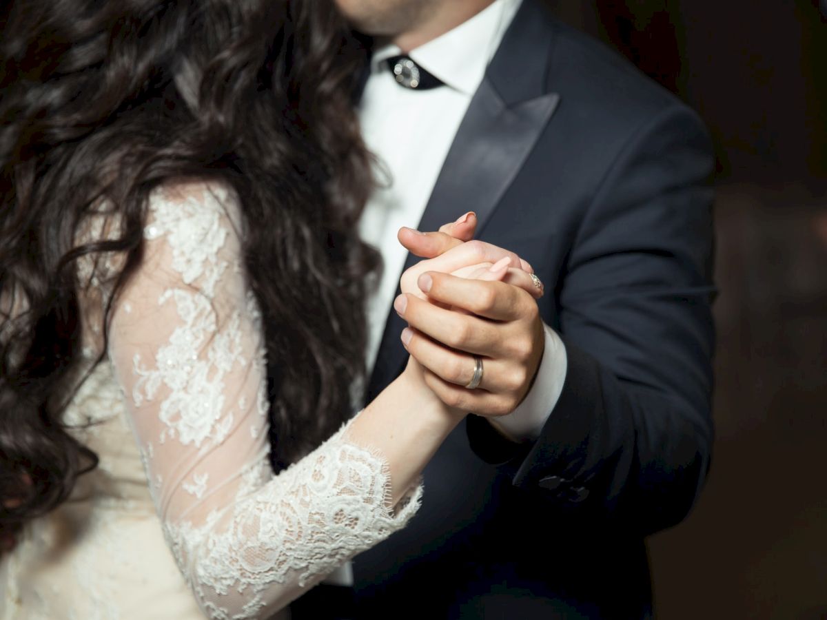 A couple is dancing closely, with their hands clasped together. The woman is wearing a white lace dress, and the man is in a dark suit and tie.