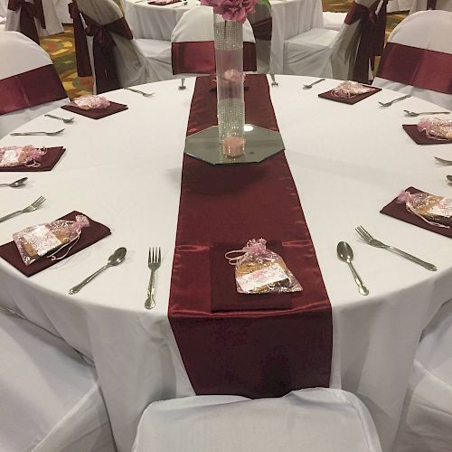 A round table set for an event with white tablecloth, maroon runner, napkins, cutlery, and a tall centerpiece with pink flowers in the middle.