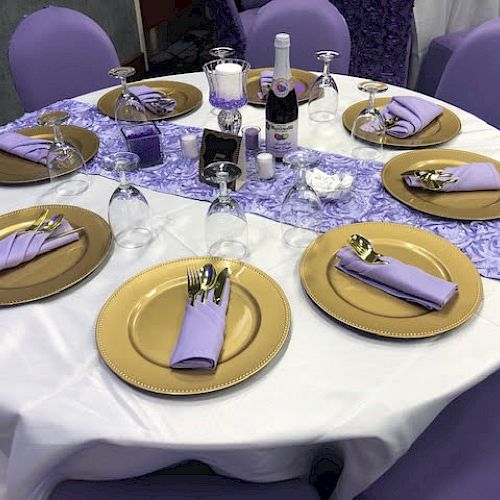 A round table set for an event with gold plates, purple napkins, utensils, and purple-themed decorations, including a bottle and candle.