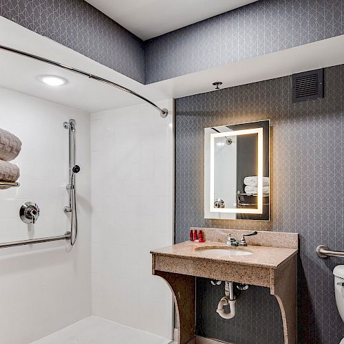 This image shows a modern bathroom with a walk-in shower, toilet, vanity with a mirror, towel rack with folded towels, and grab bars for accessibility.