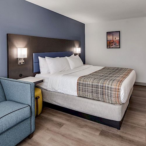 This image shows a modern hotel room with a bed, blue sofa, wooden flooring, wall decor, and a door leading to an entrance or bathroom area.