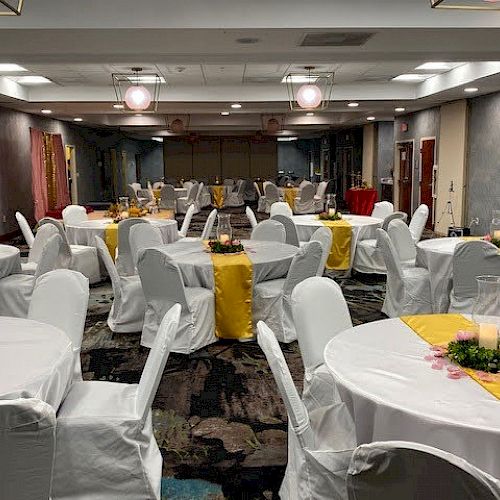 A banquet hall is set up with round tables covered in white tablecloths and yellow runners, with white chair covers, and a floral centerpiece.