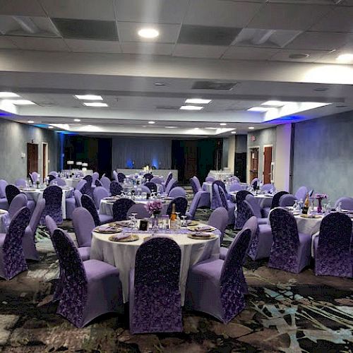 A banquet hall with multiple round tables adorned with white tablecloths and settings, with purple chair covers, prepared for an event.