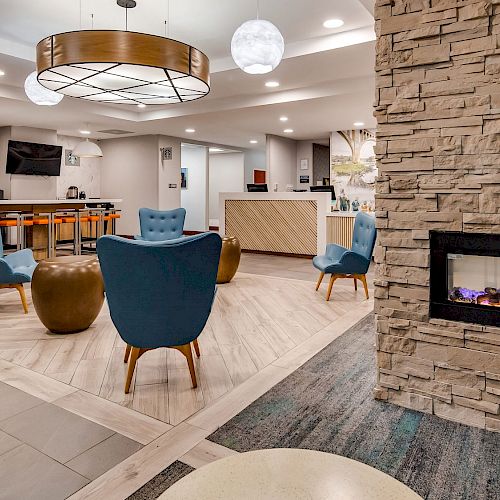 The image shows a modern lobby with stylish chairs, a stone fireplace, a reception desk, and a sitting area with a television screen.