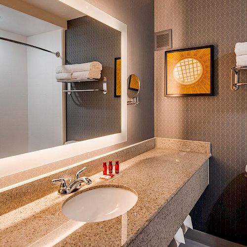 The image shows a modern bathroom with a large illuminated mirror, countertop with sink, towel rack holding towels, and a toilet, in a well-lit setting.