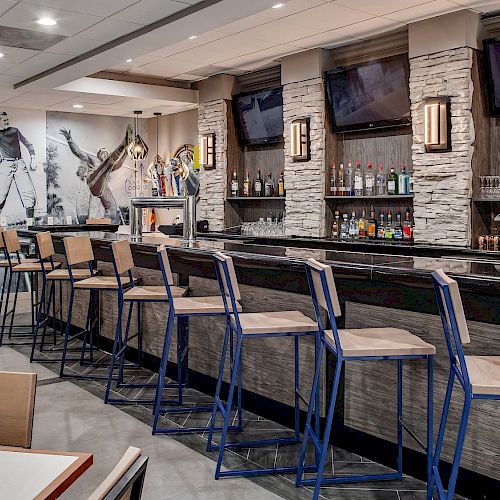 A modern bar with high chairs, a fully stocked bar counter, and a backdrop of sports-themed wall art. The seating area includes tables and chairs.