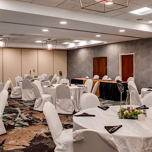 A banquet hall with round tables and chairs covered in white linens, decorated with centerpieces and black napkins, ready for an event.