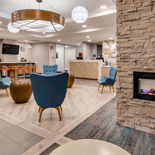A modern lounge area features blue chairs and a stone fireplace, with a reception desk and a bar counter in the background.